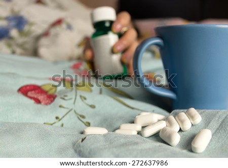 Spilled pills, blue cup on a messy bed and female hand with  medications pill bottles in the blurred background