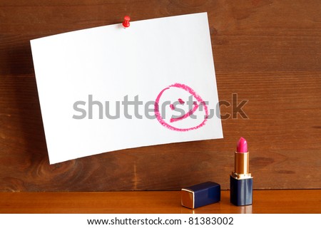 Lipstick standing near blank paper sheet with drawn smiling face. Paper is hanging on wooden wall