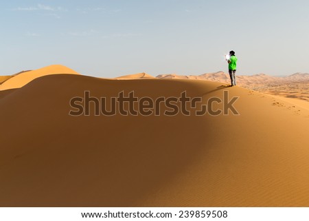 One person on a dune in the desert, holding a touch pad. Sun reflection on the screen. Landscape