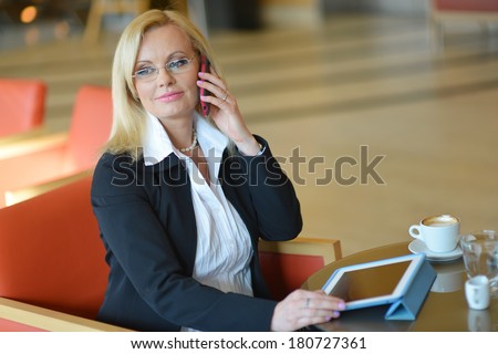 Candid photo of a attractive middle-aged blond businesswoman working at hotel lobby with a tablet pc/smartphone and drinking coffee