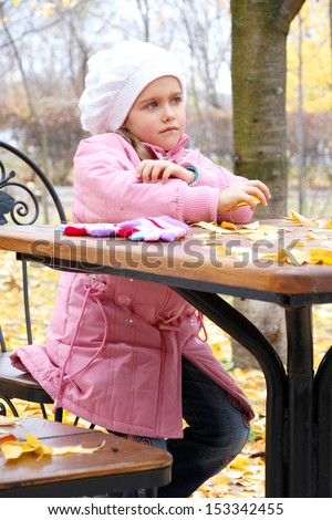 Little sad beautiful girl sitting at the table empty cafe on background of autumn park.