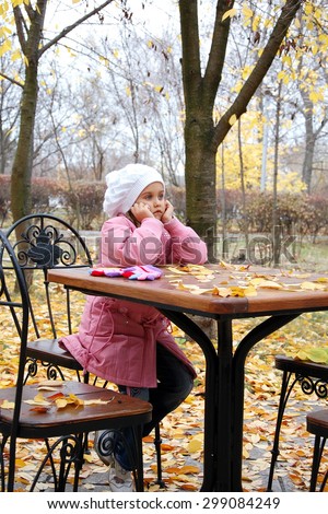 sad girl in the autumn. little girl sitting alone at a table cafe.\
girl in a pink coat was left alone. No family, no parents. Girl waiting for mum and dad. without family