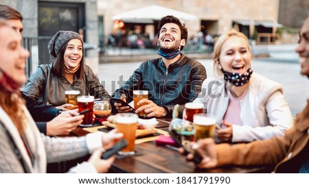 Young friends drinking beer wearing face mask - New normal lifestyle concept with people having fun together talking on happy hour at hipster brewery bar - Bright warm filter with focus on central guy