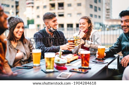 Young friends drinking beer pint with open face mask - New normal lifestyle concept with milenials having fun together talking at outside brewery bar - Warm filter with focus on left central guy