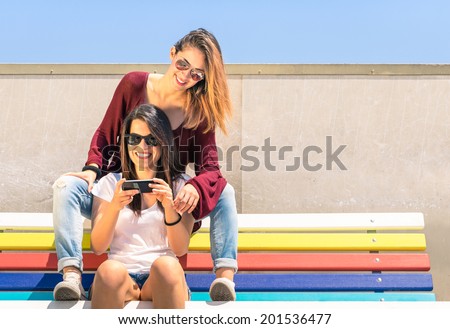 Best friends enjoying time together outdoors with smartphone - Concept of new technology with two girlfriends having fun on a multicolored bench