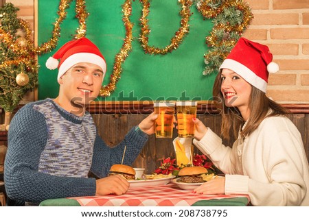 Happy couple of best friends at american restaurant with santa hats - Food and fun during Christmas holidays - Enjoying beer and hamburgers during a winter dinner