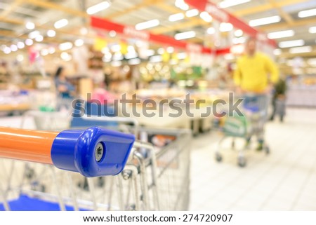 Blurred defocused grocery supermarket - Consumerism concept in period of economic crisis - Bright vivid blur of people in commercial center - Shallow depth of field with focus on shopping cart edge