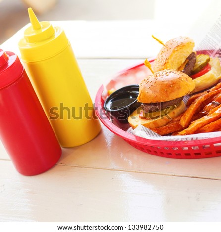 Plate of mini burgers and fries with tomato ketchup and mustard sauce bottle on table. Vertical shot.