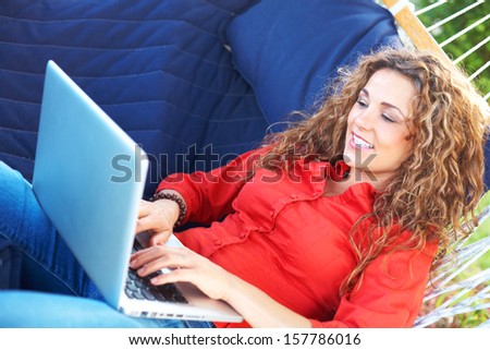 happy beautiful female laying in hammock typing on laptop looking at screen. Horizontal, color image, young woman on vacation relaxing in hammock with lap top.