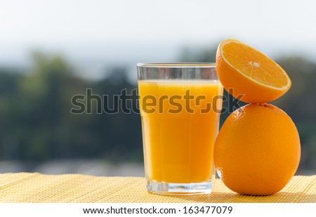 A cut fresh orange on top of another orange citrus fruit next to a glass of orange juice placed to the right side of the shot with blurry green trees in the left side of the shot