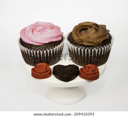 Chocolate and raspberry frosted chocolate cupcakes with brown and red chocolate candy hearts and flowers on a white ceramic pedestal against a white background