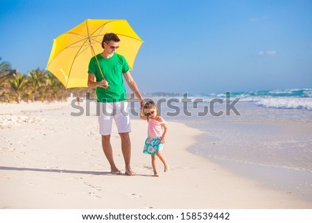 Young father and his little daughter walking under a yellow umbrella on white sand beach