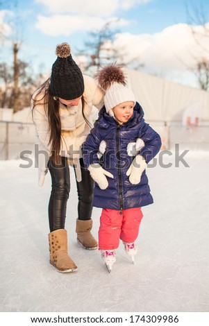 Young mother teaching her little daughter to skate on ice-rink