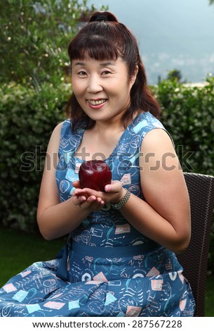 Beautiful Japanese lady outdoor/Happy Japanese lady/A photo of a beautiful middle aged Japanese lady presenting a ripe apple in a lush garden with lake Como, Italy in the background. 3/4 portrait.