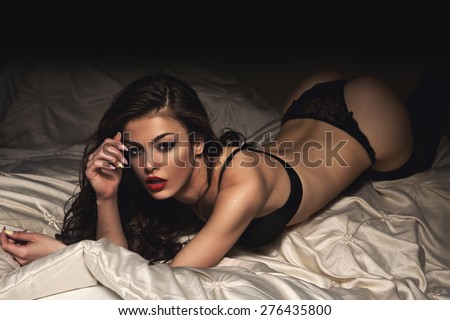 Sexy beautiful brunette woman lying in bed in sensual black lingerie, looking at camera.