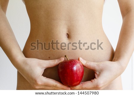 Young woman holding a red apple against her flat stomach. Concept of a good physical shape in a connection to a healthy nutrition / Adame and Eve story
