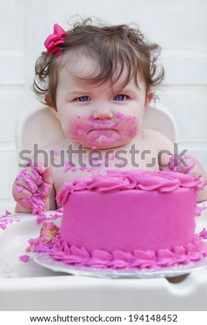 Closeup of a baby girl eating her first birthday cake with bright pink frosting looking into camera./Baby girl eating first birthday cake with pink frosting and bow in her hair