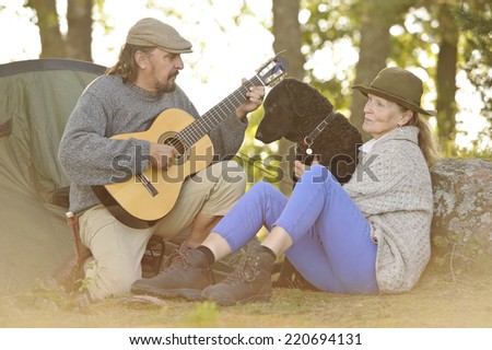 Senior couple enjoying some music outside their tent in evening light. Man plays guitar. They have their pet curly coated retriever with them. Natural lens flare.