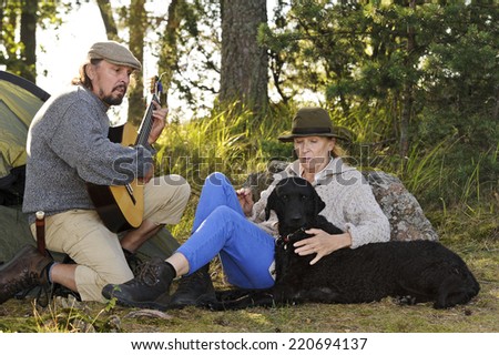 Senior couple enjoying some music outside their tent in evening light. Man plays guitar and they sing together. They have their pet curly coated retriever with them
