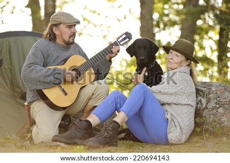 Senior couple enjoying some music outside their tent in evening light. Man plays guitar. They have their pet curly coated retriever with them. Natural lens flare