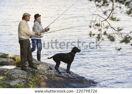 Senior couple fishing from the lake shore. Curly haired retriever pet looks on
