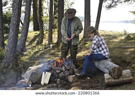 Senior couple sit by a camp fire. Man is paring sticks for grilling with. In the background there are trees, a lake, and in the distance a bridge can be seen