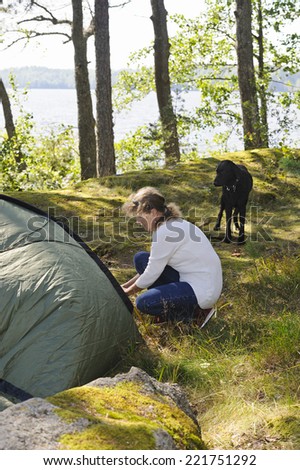 Senior woman pitches a tent in the forest. A curly haired retriever dog watches. There's a lake in the background