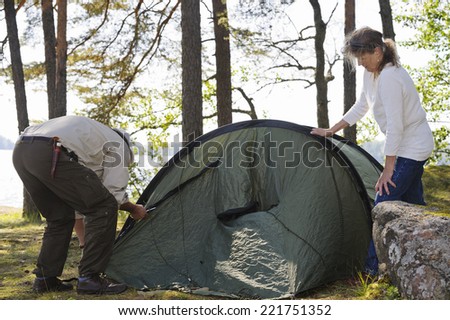 Senior couple pitch a tent together in the forest. There\'s a lake in the background.