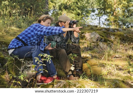 Senior couple have a tea or coffee break in the forest while hiking. They use  binoculars to check out the landscape. Focus is on the woman