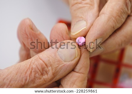 Elderly hands hold a pink tablet between the fingers. A pill organizer box is visible behind his hands.