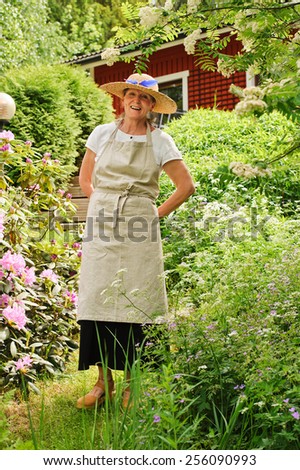 Senior woman standing in her garden, She is surrounded by bushes. There\'s a red house partly seen in the background. She\'s wearing an apron