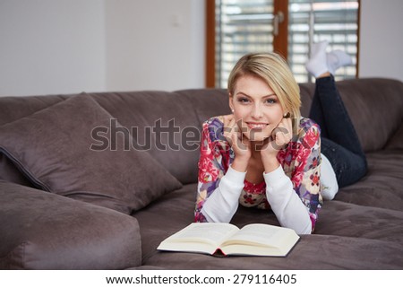 Pretty young woman enjoying reading a book at home lying on the sofa smiling in pleasure in casual clothing