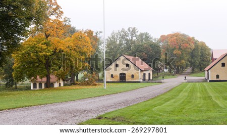 View into a garden park and a few yellow buildings. Green lawn and and some mist in the air. Fallen branch on the ground. Part of farmhouses.