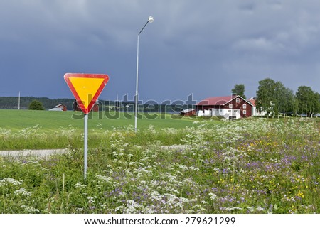 Give way at the entry to the highway. Farmland, flowers and showers. Buildings in the background.