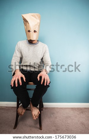 A Man with a paper bag over his head is sitting on a chair against a blue wall