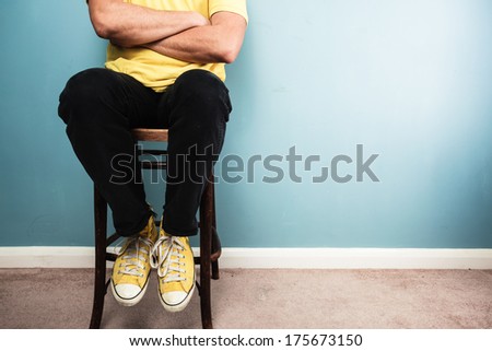Below the neck shot of a young man sitting on a chair against a blue wall