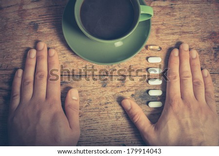 A man is resting his hands on a table with a cup of coffee and some pills