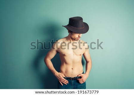 A sexy shirtless cowboy is posing by a blue wall