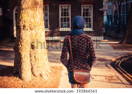 A young woman is walking past a tree and a townhouse in the city on a sunny day