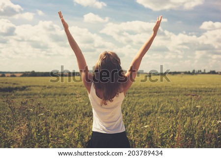 A young woman is standing in a wheat field in summer and is raising her arms up to the sky