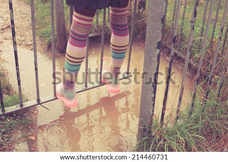 A young woman wearing rain boots is standing by a puddle and a gate