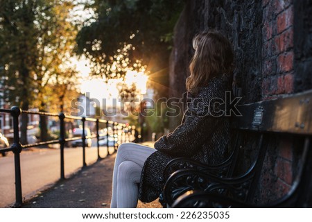 A young woman is sitting on a bench at sunset on an autumn day in the city