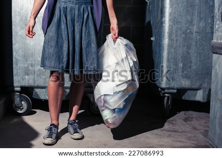 A young woman is standing next to some large bins with a bag of rubbish
