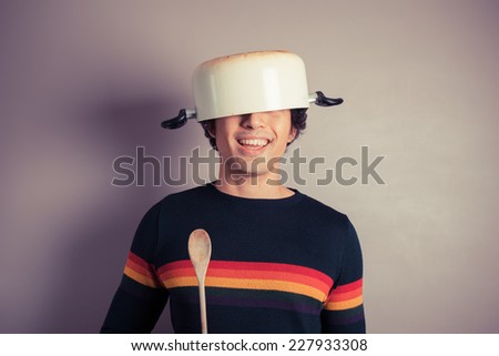 A silly young man is wearing a cooking pot on his had and is holding a wooden spoon