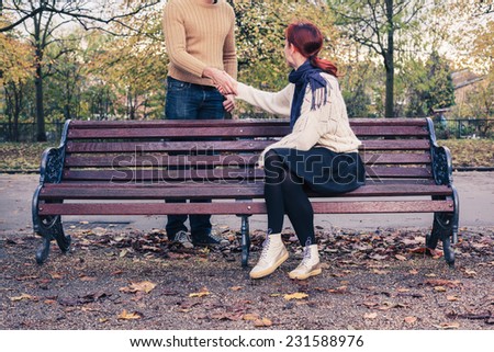 A young woman sitting on a park bench is meeting a man in the park and they are shaking hands
