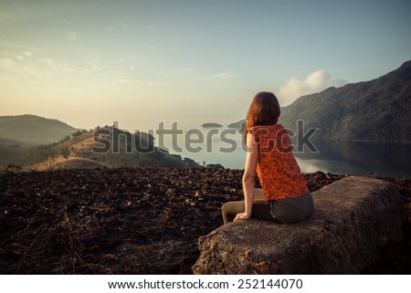 A young woman is sitting on an unusual rock on a mountain overlooking a bay at sunrise in a tropical climate