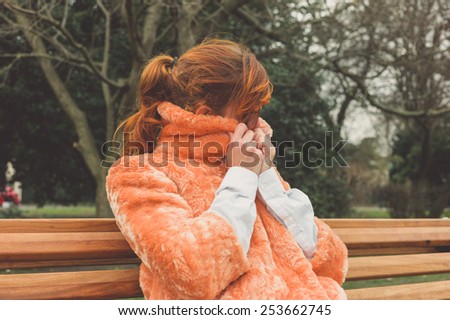 A young woman is sitting on a park bench and is wrapping up in her coat on a winter\'s day