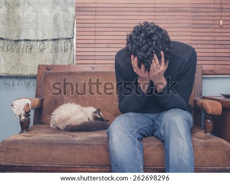A sad and depressed young man is sitting on a sofa with a cat and his head buried in his hands
