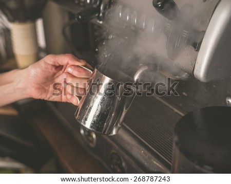A female hand is placing a cup under the froth element of a professional coffee machine which causes steam to rise from the liquid in the cup