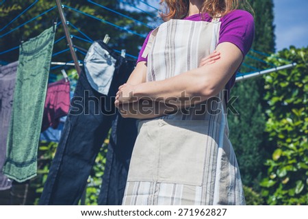 A young woman wearing an apron is standing by her laundry drying on a clothes line in the garden
